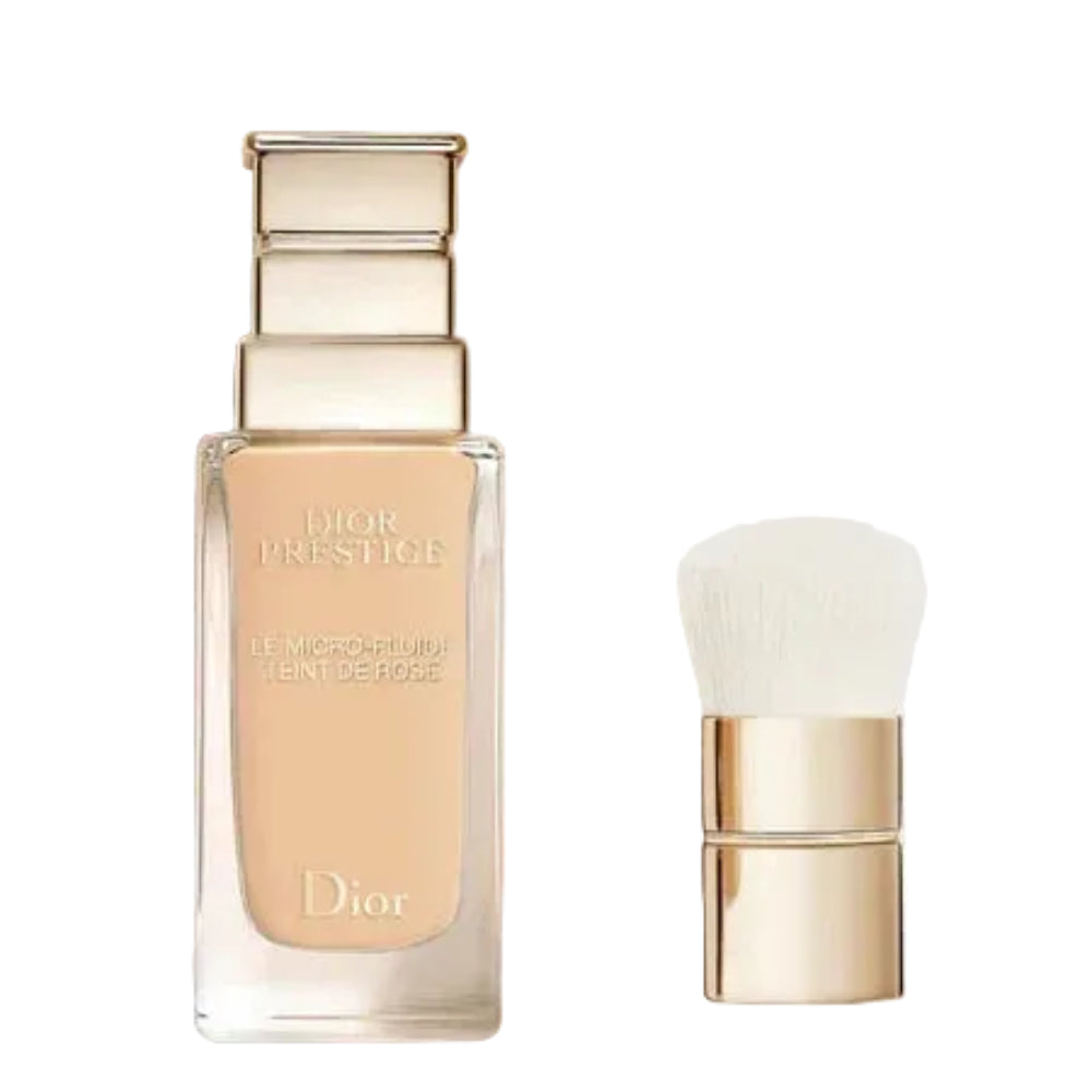Dior Prestige Le Micro-Fluidisant Tint de Rose foundation bottle in a clear glass bottle showing the beige liquid, next to a matching gold and white applicator brush, on a light gray background.